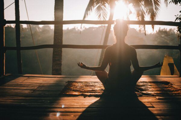 A woman meditating on a wooden deck before palm trees, the sun flaring in the distance, rendering everything else, in comparison, dark
